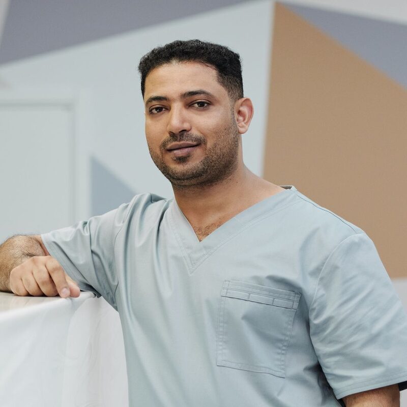 Sam Haddad in light blue scrubs leaning on the front desk and smiling