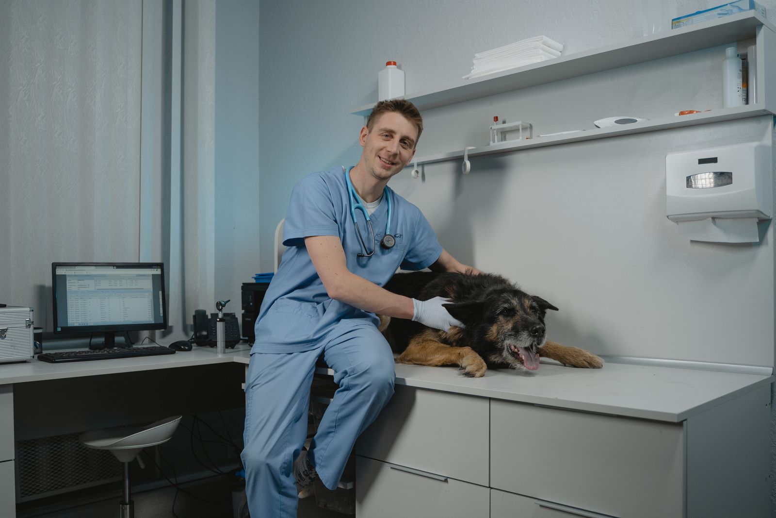 A smiling veterinarian in blue scrubs sitting on the exam counter with a black dog